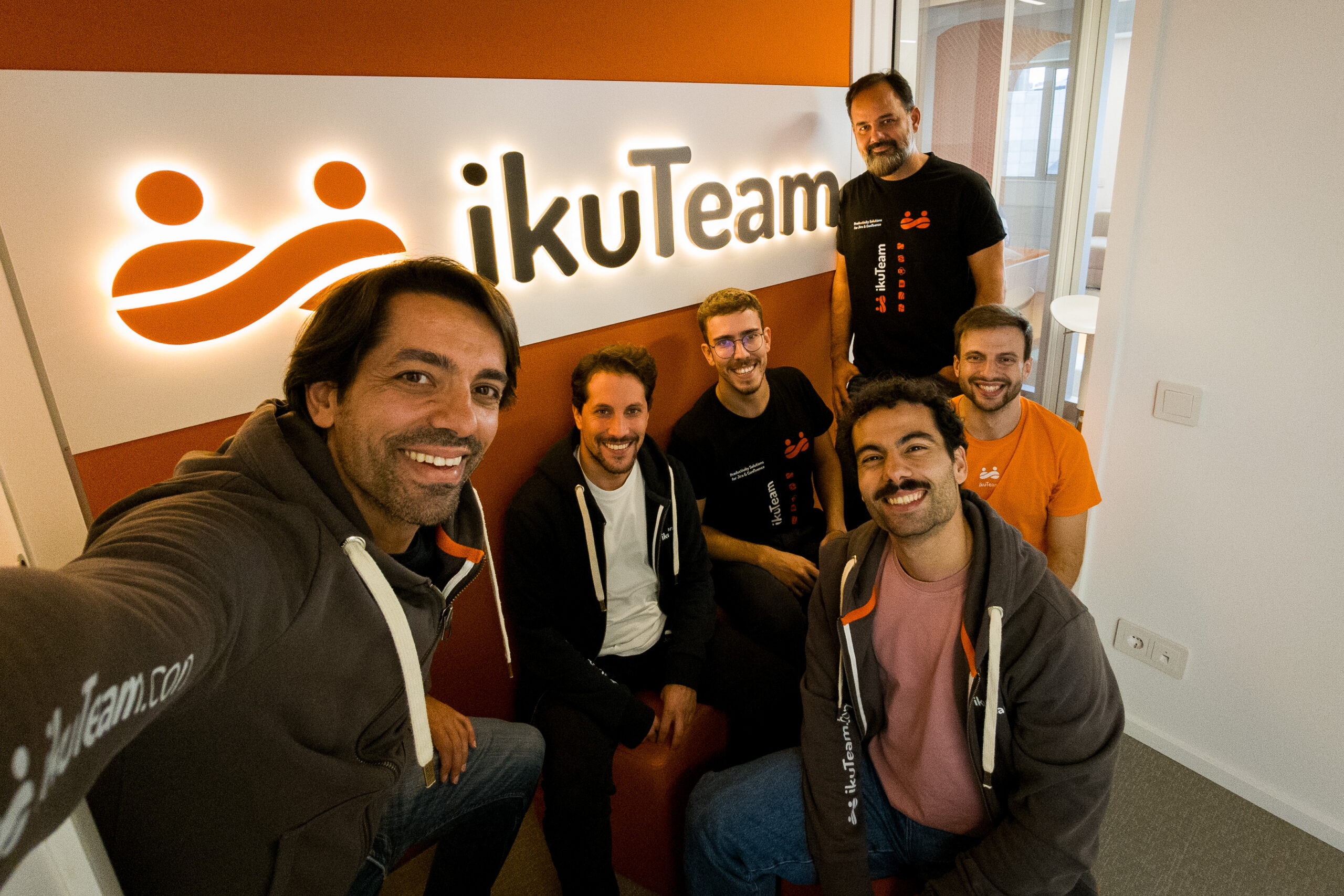 Photo of the ikuTeam team at their office, standing in front of the company logo.